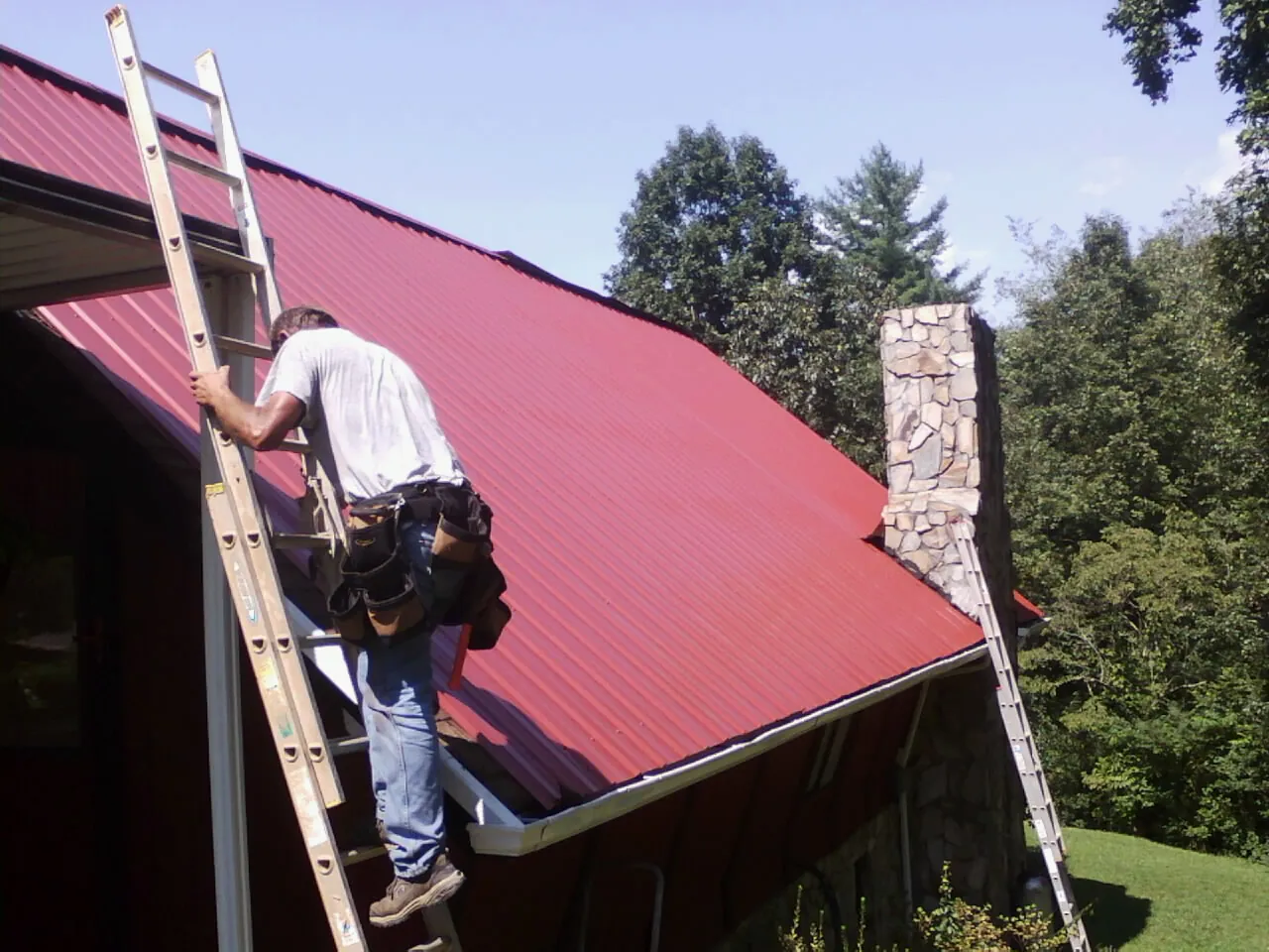Roofer on a ladder beside a red metal roof and stone chimney — Soffit & Fascia Repair by Roofing Brothers in San Marcos